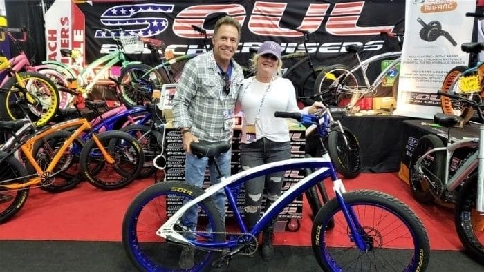 Two people standing next to a blue EBIKES at a trade show, seeking SOULFAST Support.
