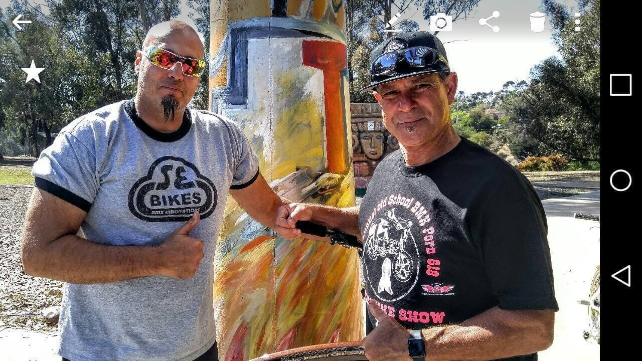 Two men posing for a photo with one man pointing towards the SOULFAST EBIKES logo on his shirt while holding a bike handle.