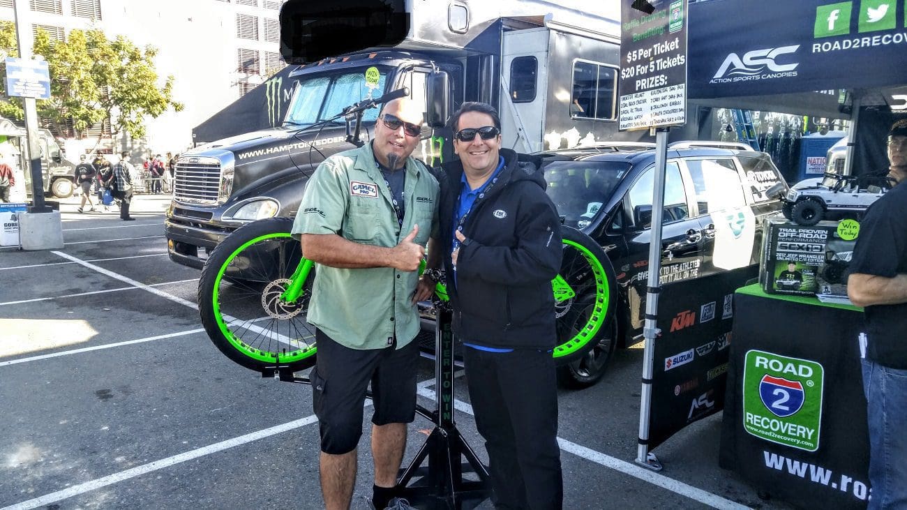 Two men smiling for the camera at an outdoor event with a SOULFAST EBIKES bike and promotional banners in the background.