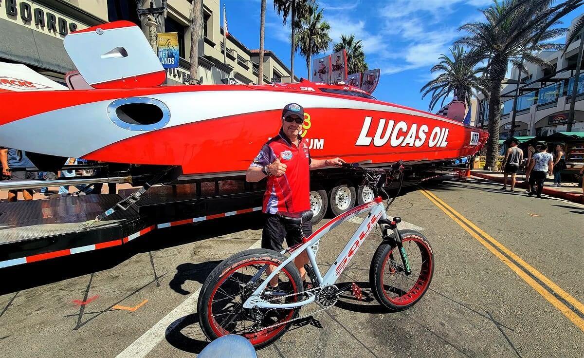 Man posing with bike beside racing boat.