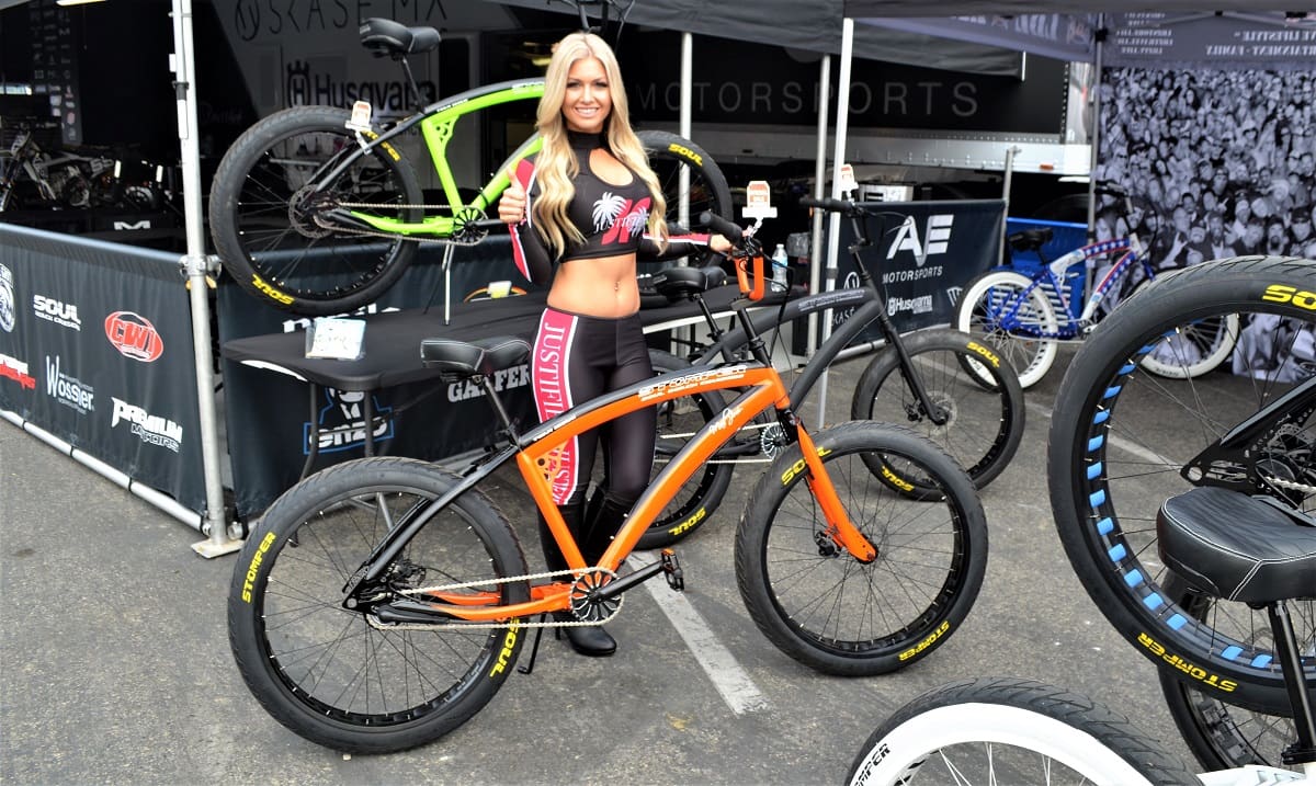Woman posing with orange bicycle at event.