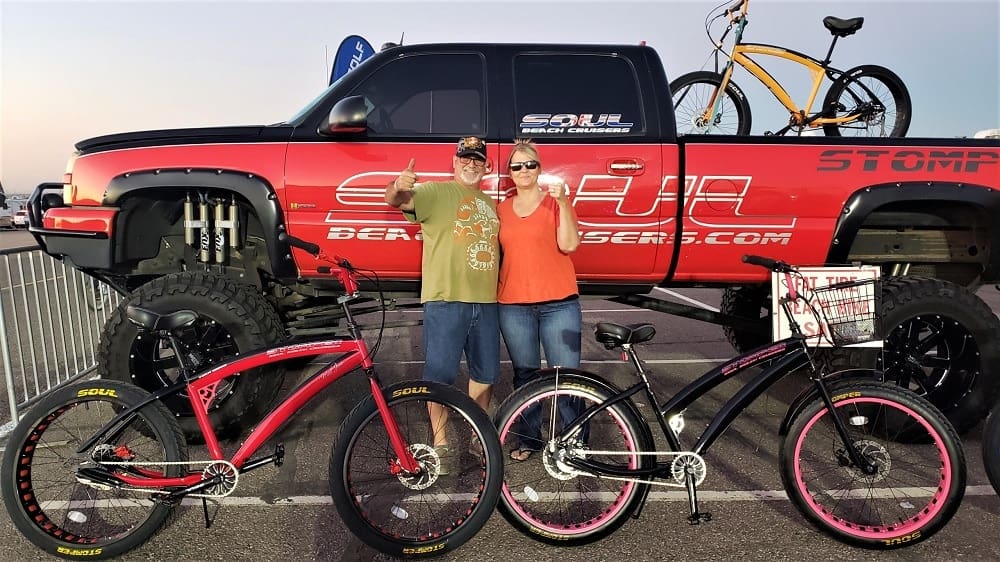 Couple standing with bicycles, truck in background.