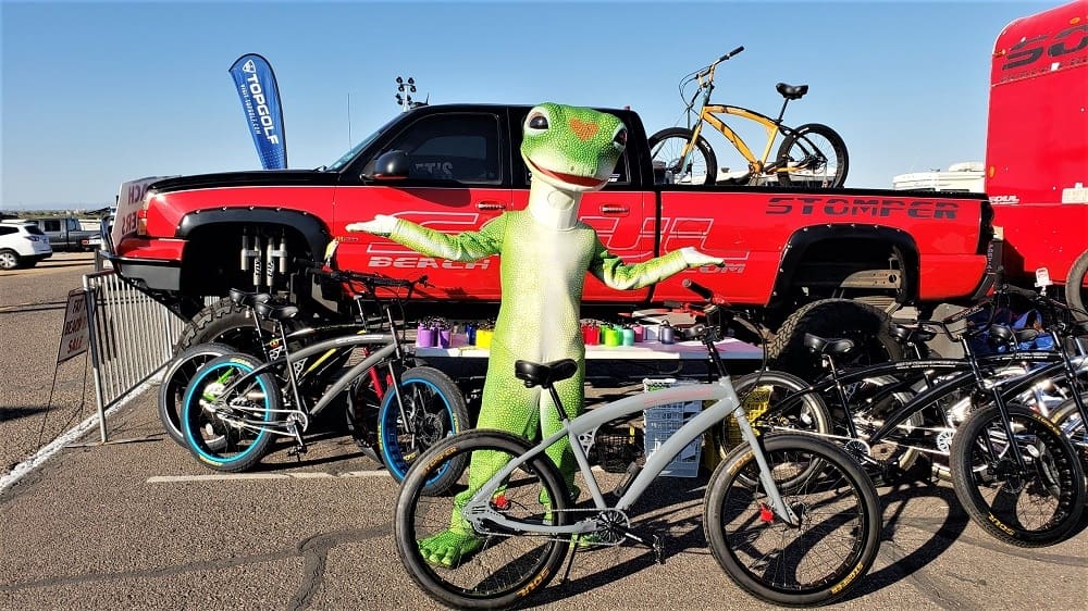 Lizard mascot posing with bicycles and truck.