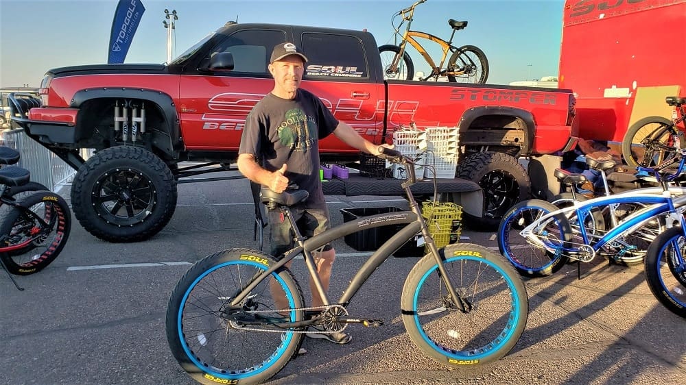 Man posing with bicycle at outdoor event.