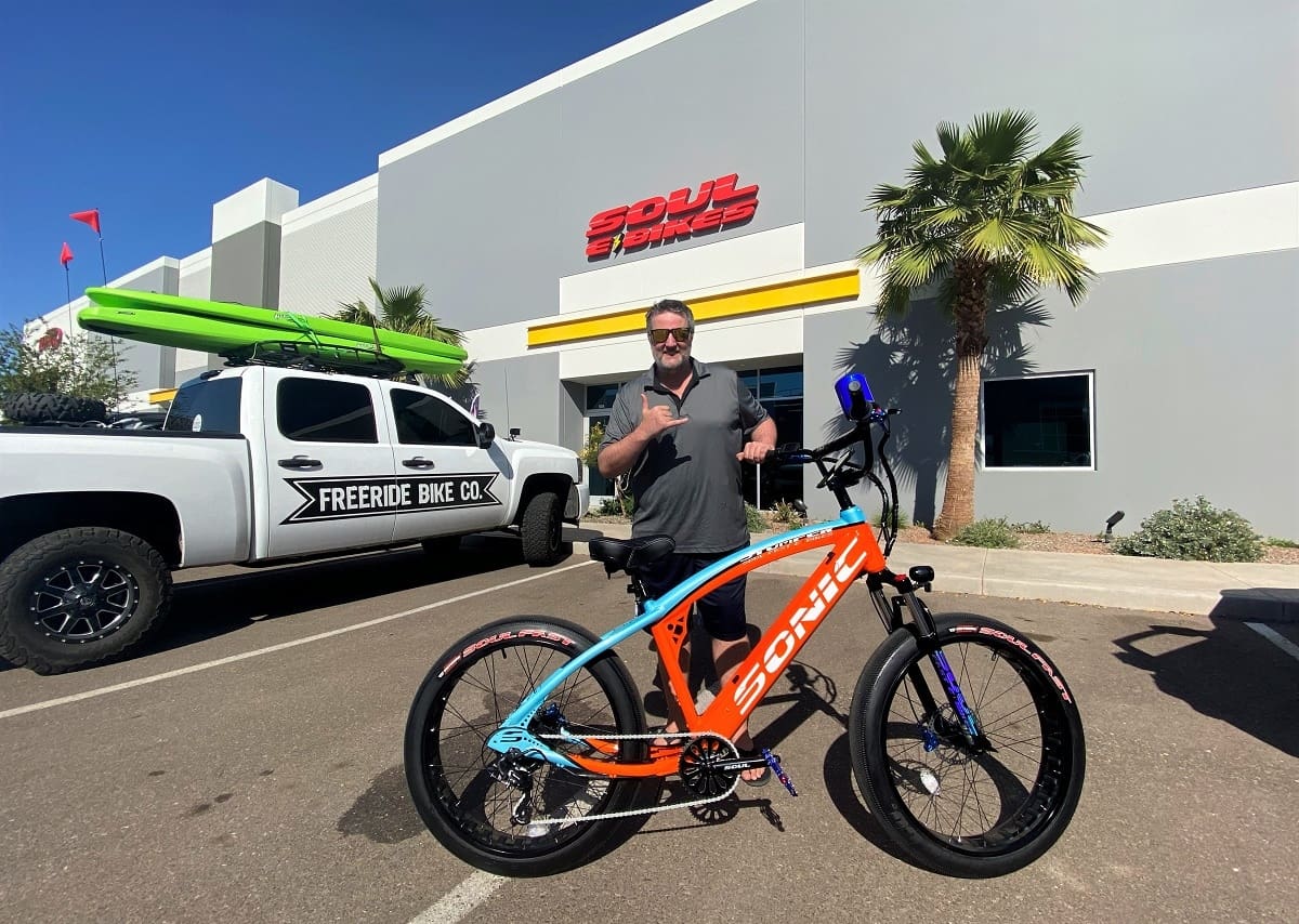 Man posing with orange electric bicycle outside store.