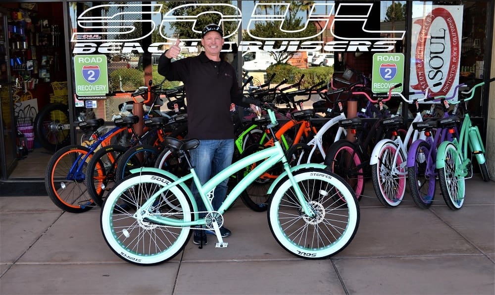 Man posing with colorful cruiser bicycles outside shop.