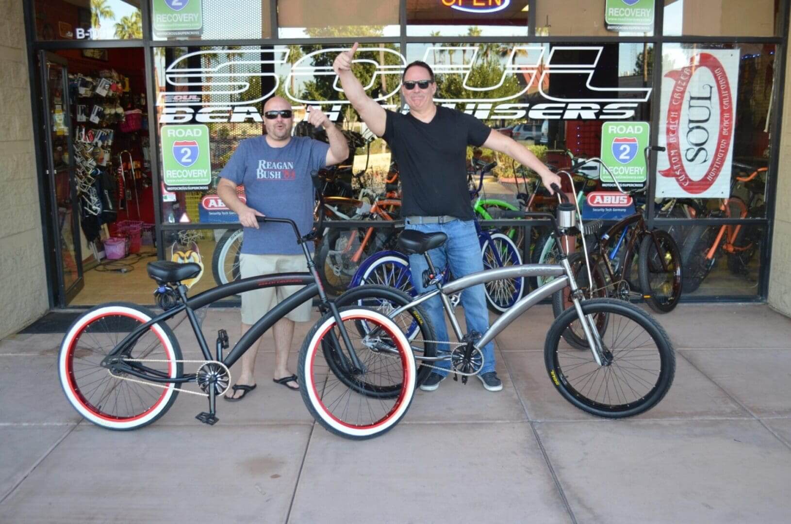 Two men posing with bicycles outside shop.