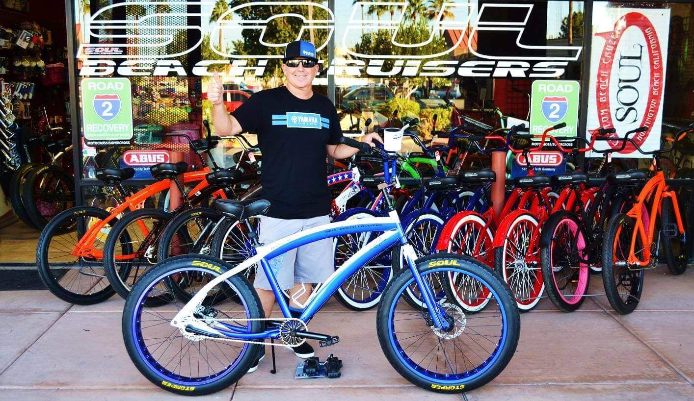 Man posing with blue bicycle outside shop.