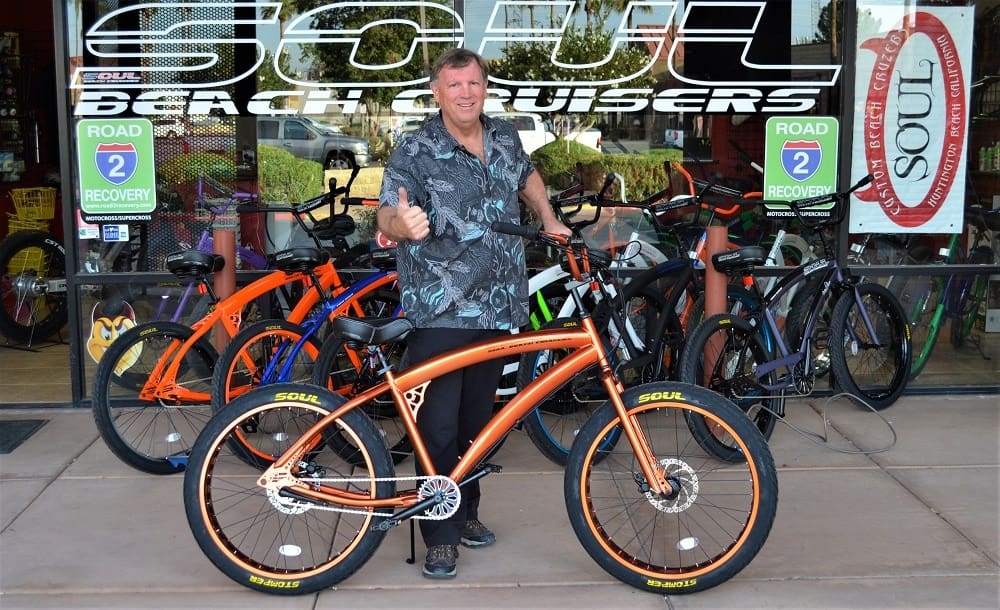 Man posing with orange bicycle outside shop.