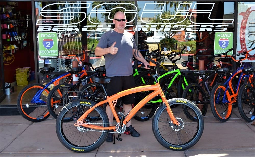 Man with orange bike outside bicycle shop.