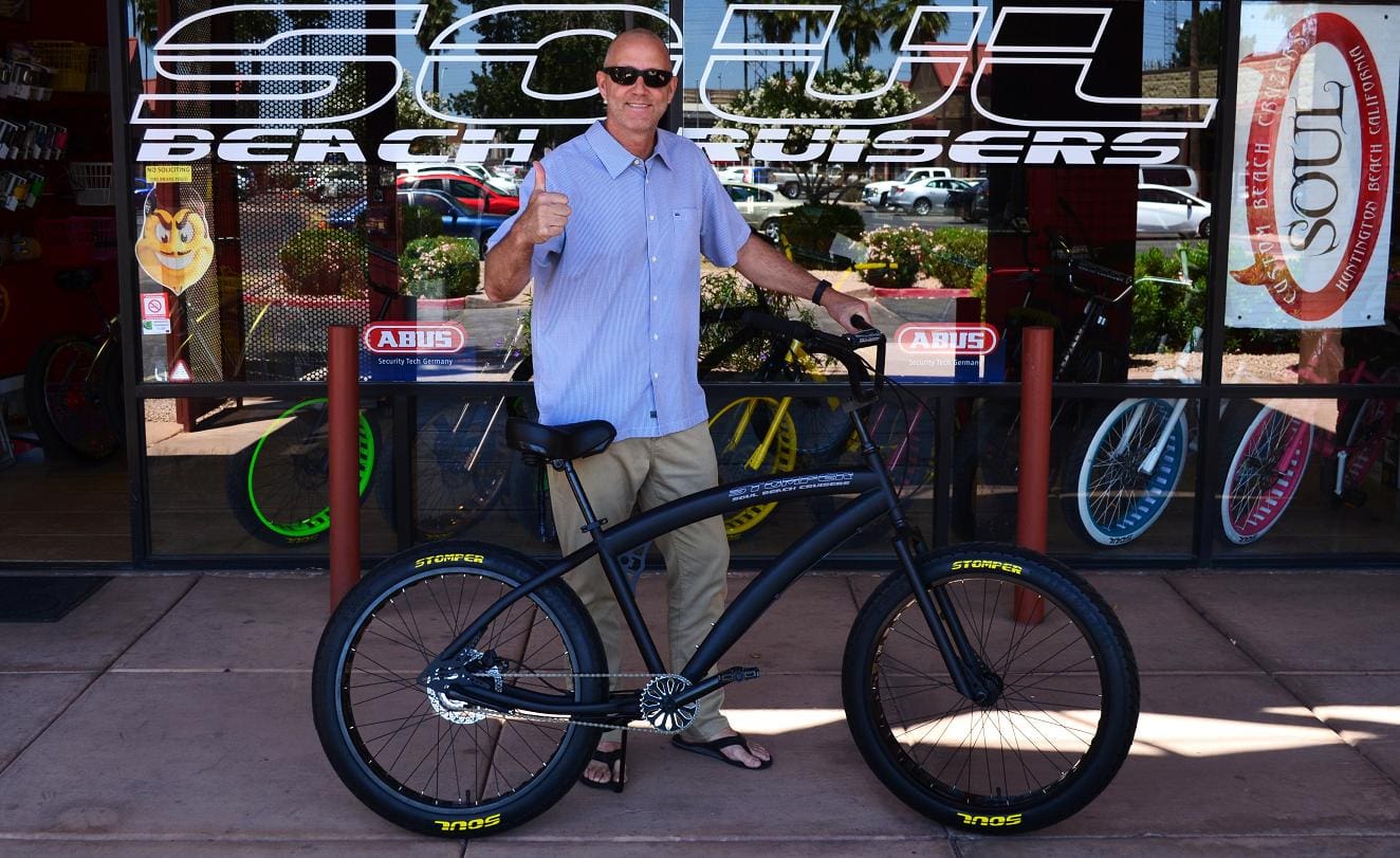 Man standing with a black bicycle outside shop.