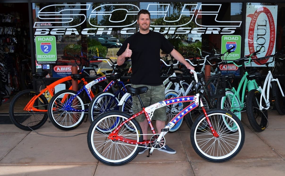 Man posing with colorful bicycles outside shop.