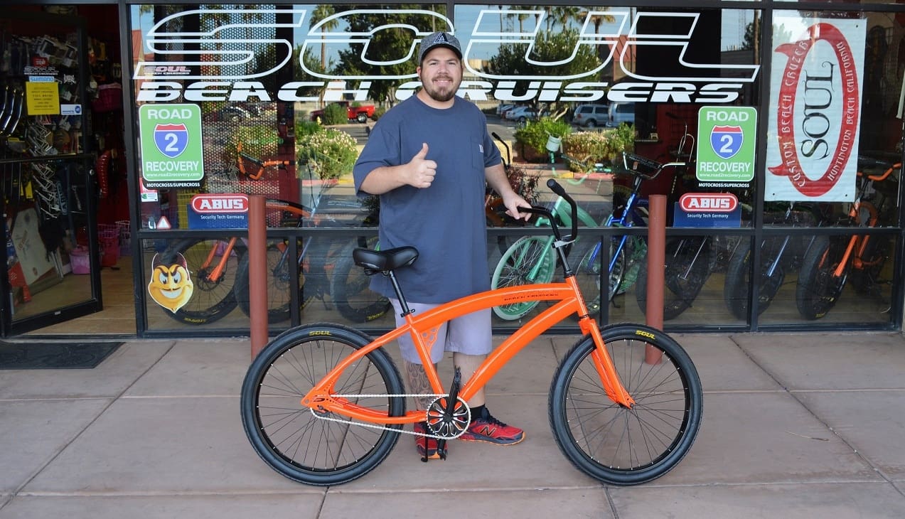 Man with orange bicycle outside bike shop.