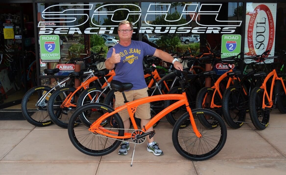 Man posing with orange bicycle outside shop.
