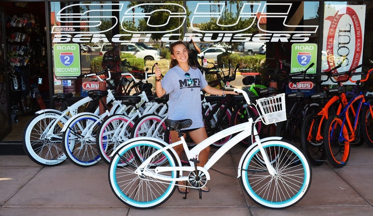 Woman smiling with white bike outside shop.