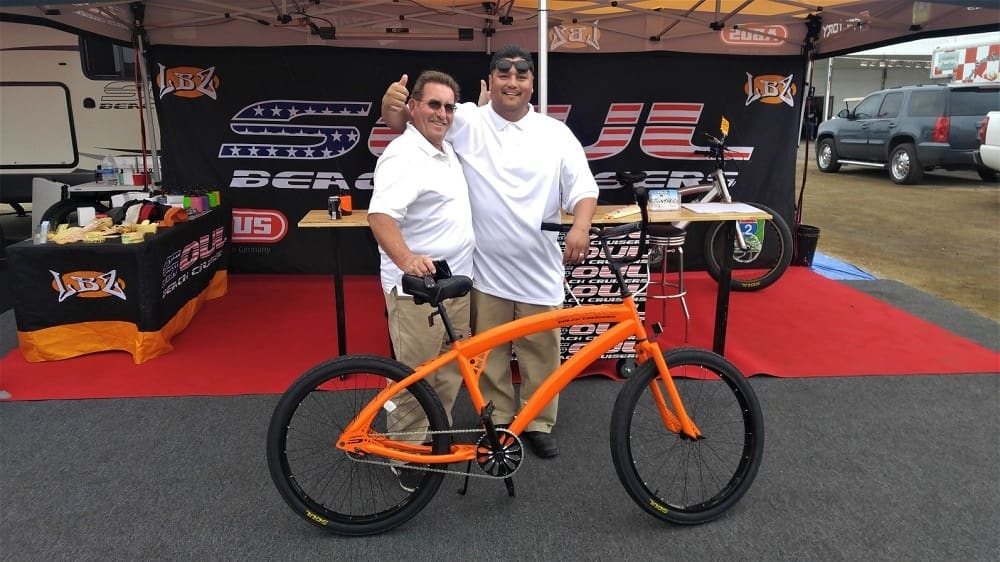 Two men posing with an orange bicycle.