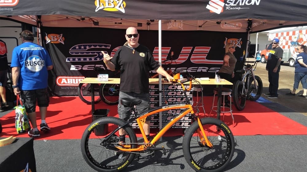 Man posing with orange bicycle at event.