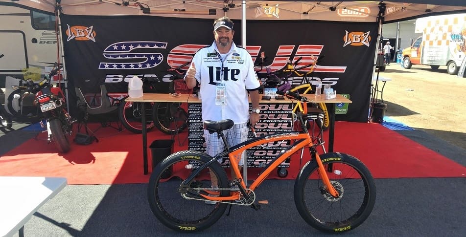 Man posing with orange bicycle at event.