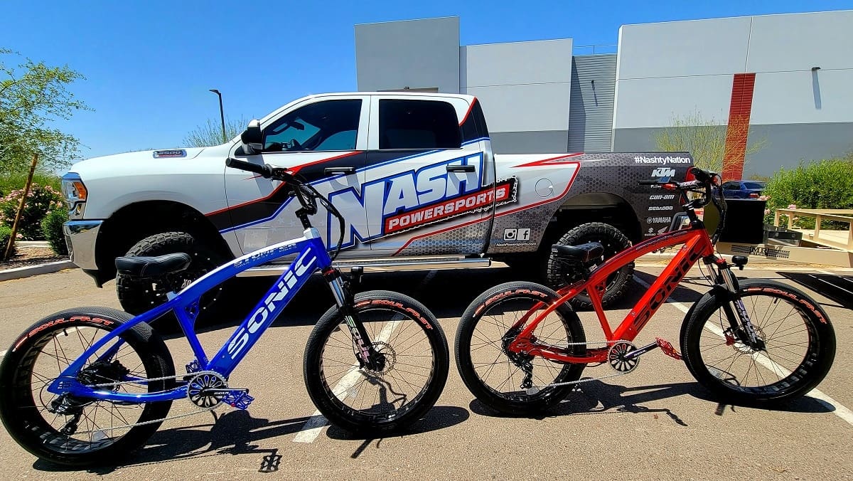 Two electric bikes beside branded pickup truck.