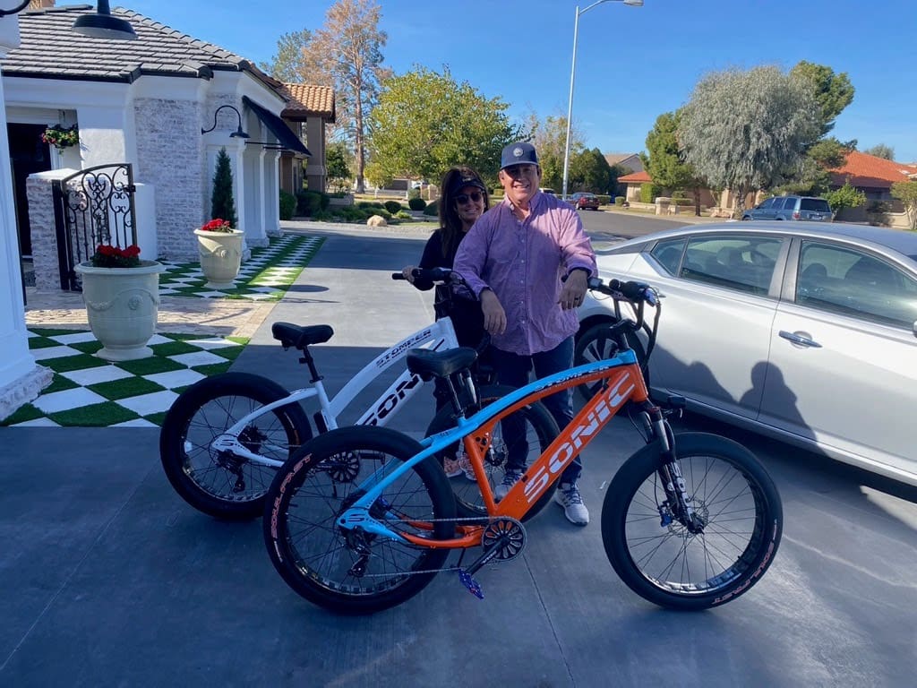 Couple posing with electric bicycles outside house.