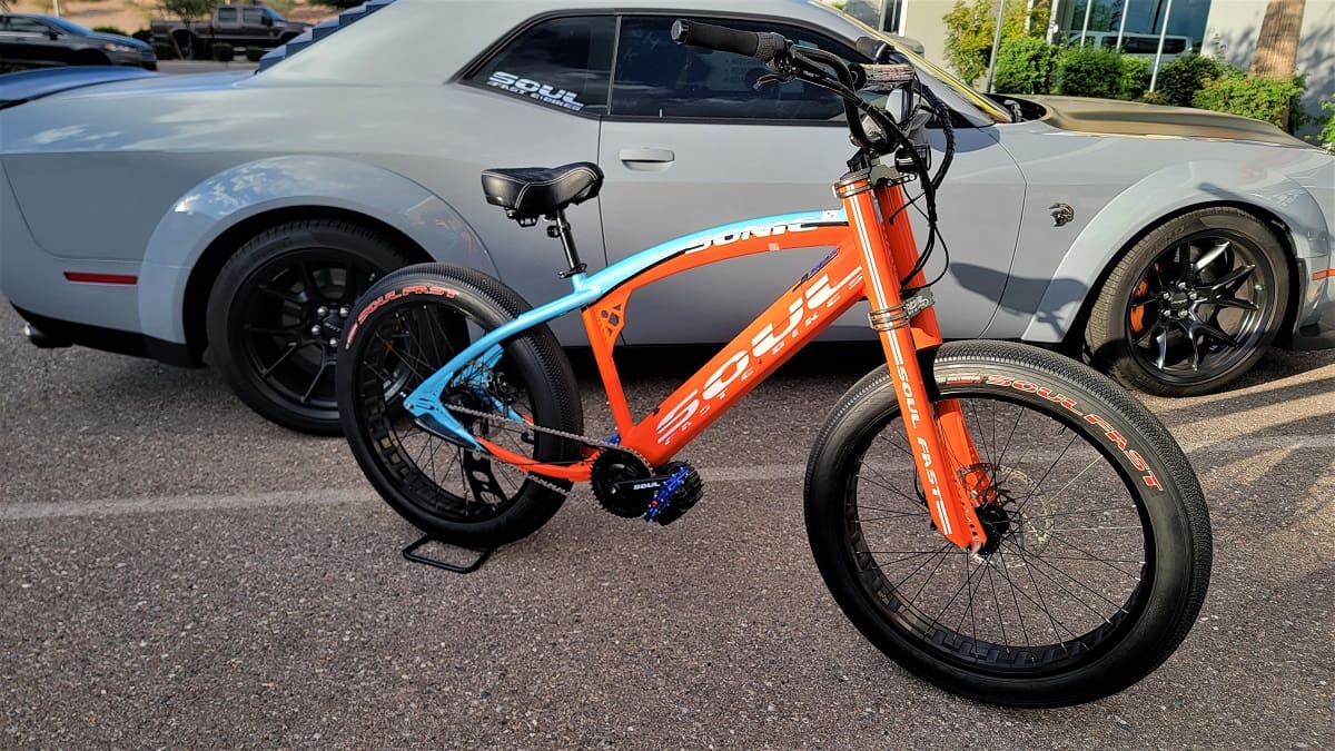 Orange and blue bicycle beside gray car.
