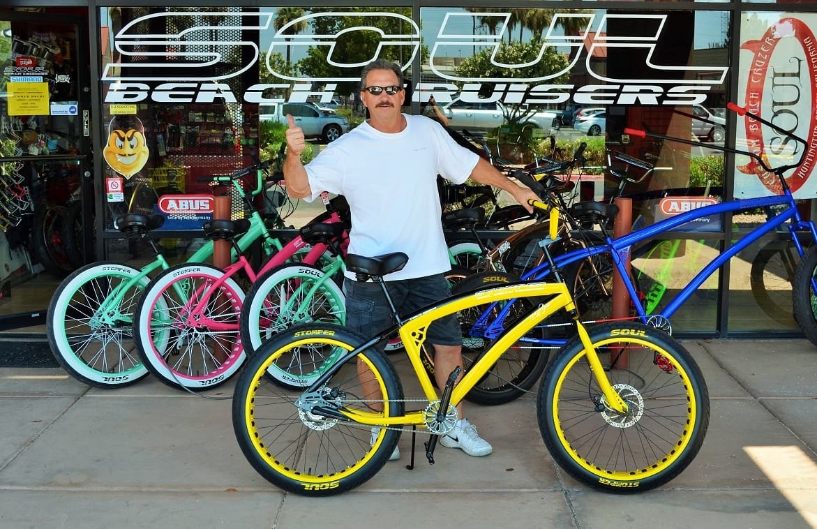 Man posing with colorful bicycles outside shop.