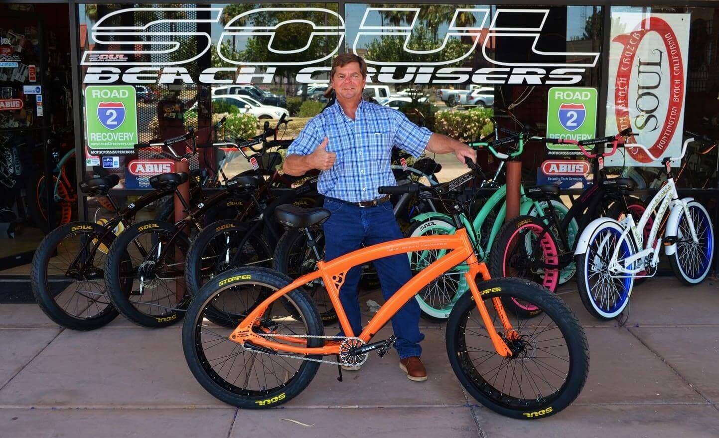 Man posing with orange bicycle outside shop.