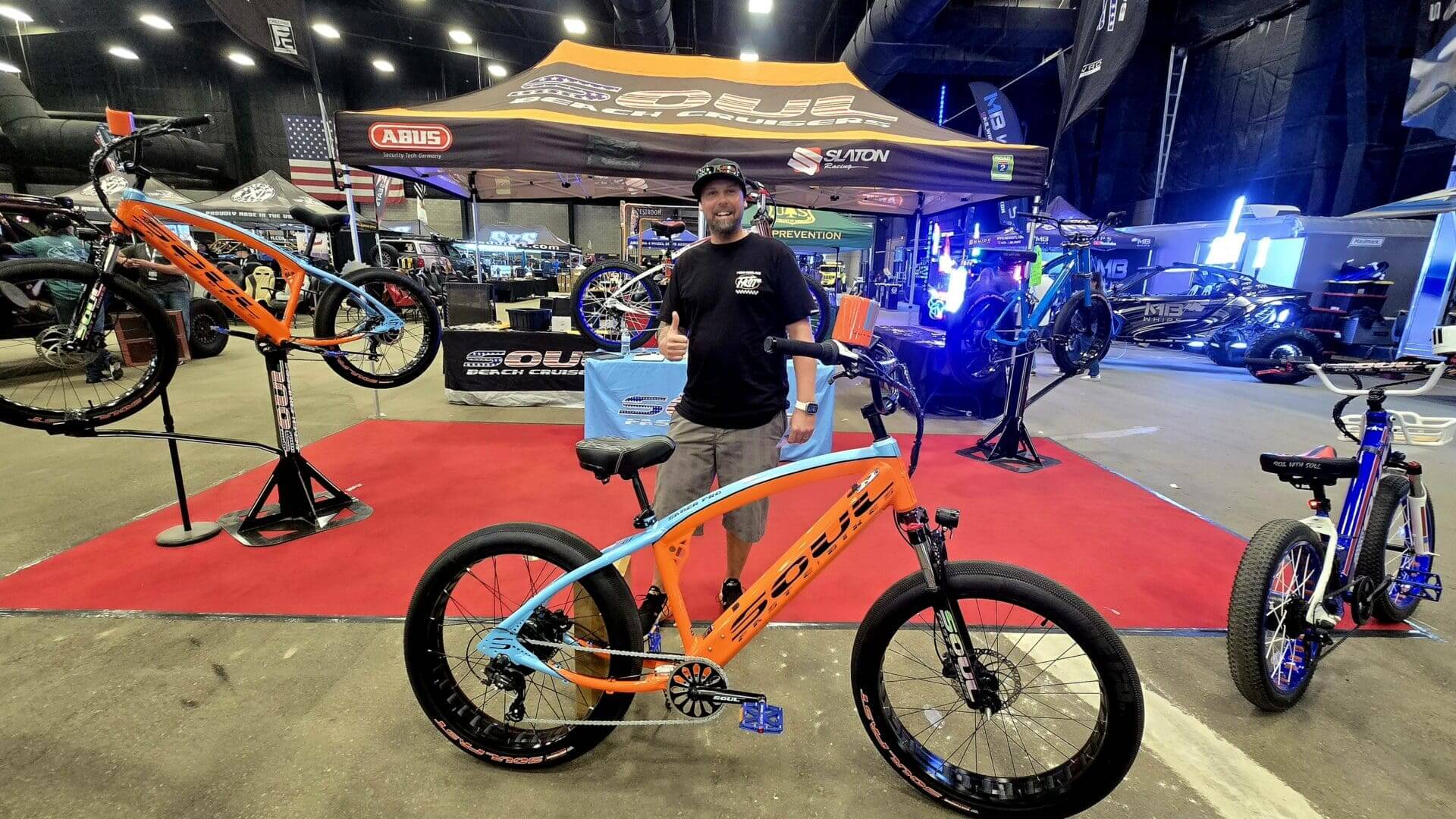 Man posing with orange bicycle at exhibition.